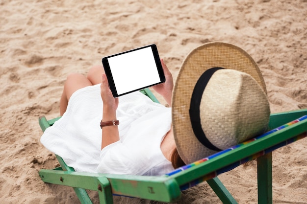 Imagen de maqueta de una mujer sosteniendo una tablet pc negra con pantalla de escritorio en blanco mientras está acostada en una silla de playa