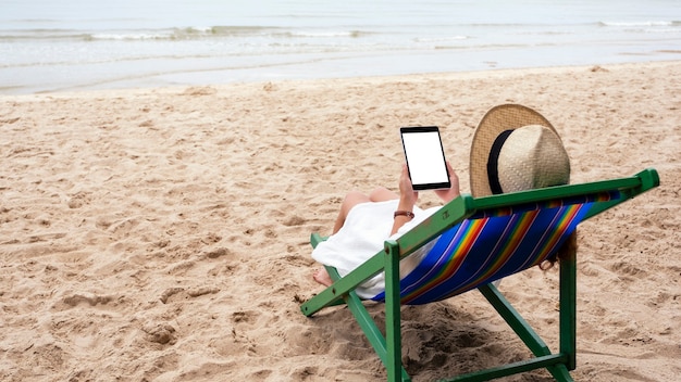 Imagen de maqueta de una mujer sosteniendo una tablet pc negra con pantalla de escritorio en blanco mientras está acostada en una silla de playa