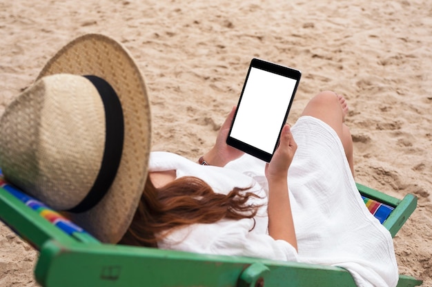 Imagen de maqueta de una mujer sosteniendo una tablet pc negra con pantalla de escritorio en blanco mientras está acostada en una silla de playa