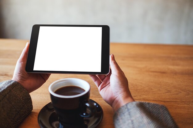 Imagen de maqueta de mujer sosteniendo tablet pc negro con pantalla en blanco en blanco con taza de café en la mesa de madera