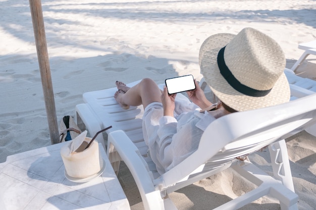 Imagen de maqueta de una mujer sosteniendo un teléfono móvil blanco con pantalla de escritorio en blanco mientras se acuesta en una silla de playa en la playa