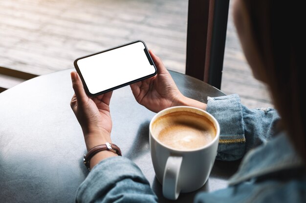 Imagen de maqueta de una mujer sosteniendo un teléfono móvil negro con pantalla en blanco con una taza de café en la mesa de madera