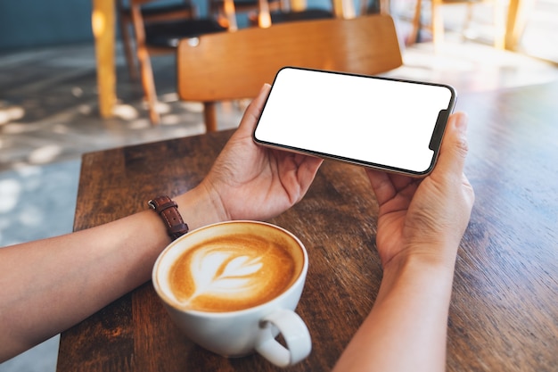 Imagen de maqueta de una mujer sosteniendo un teléfono móvil negro con pantalla en blanco con una taza de café en la mesa de madera