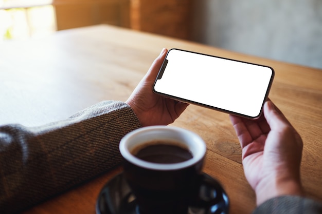 Imagen de maqueta de una mujer sosteniendo un teléfono móvil negro con pantalla en blanco con una taza de café en la mesa de madera