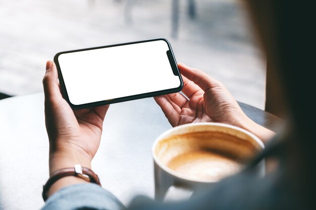 Imagen de maqueta de una mujer sosteniendo un teléfono móvil negro con pantalla en blanco con una taza de café sobre la mesa