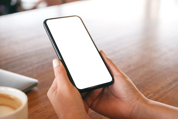 Imagen de maqueta de una mujer sosteniendo un teléfono móvil negro con pantalla de escritorio en blanco con una computadora portátil y una taza de café sobre la mesa