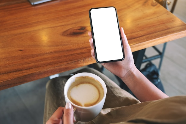 Imagen de maqueta de una mujer sosteniendo un teléfono móvil negro con pantalla de escritorio en blanco mientras bebe café