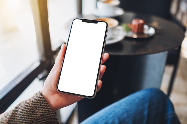 Foto imagen de maqueta de una mujer sosteniendo un teléfono móvil negro con pantalla de escritorio en blanco mientras está sentado en la cafetería