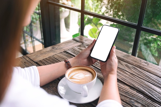 Imagen de maqueta de una mujer sosteniendo un teléfono móvil negro con pantalla de escritorio en blanco con una taza de café sobre la mesa