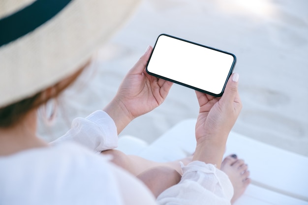 Imagen de maqueta de una mujer sosteniendo y usando un teléfono móvil blanco con pantalla de escritorio en blanco mientras está sentado en una silla de playa en la playa