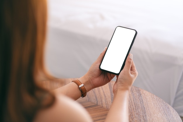 Imagen de maqueta de una mujer sosteniendo y usando un teléfono móvil con pantalla en blanco mientras está sentada junto a la cama
