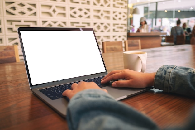 Imagen de maqueta de una mujer usando y escribiendo en una computadora portátil con pantalla de escritorio en blanco en blanco con una taza de café en la mesa de madera en la oficina