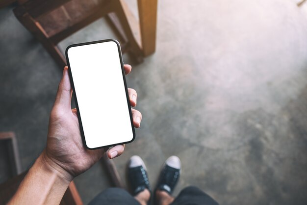 Imagen de maqueta de vista superior de un hombre de pie y sosteniendo un teléfono móvil negro con pantalla en blanco en blanco