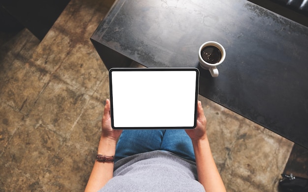 Imagen de maqueta de vista superior de una mujer que sostiene una tableta digital con una pantalla de escritorio blanca en blanco en un café