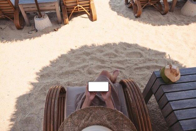 Imagen de la maqueta de la vista superior de una mujer que sostiene un teléfono móvil con una pantalla de escritorio en blanco mientras está sentada en la playa