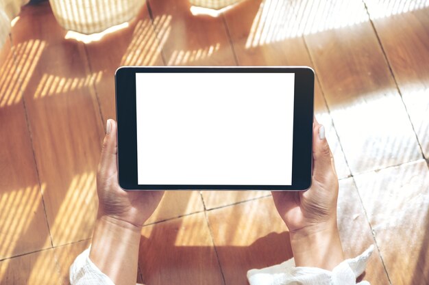 Imagen de la maqueta de la vista superior de una mujer sosteniendo una tablet pc negra con una pantalla de escritorio blanca en blanco horizontalmente mientras se acuesta en el piso de madera