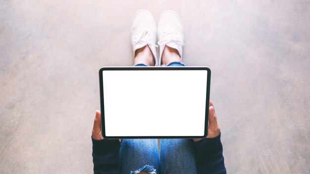 Imagen de maqueta de vista superior de una mujer sosteniendo tablet pc negro con pantalla en blanco mientras está sentada en el piso