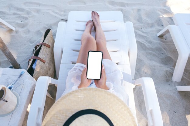 Imagen de maqueta de vista superior de una mujer sosteniendo un teléfono móvil blanco con pantalla de escritorio en blanco mientras se acuesta en una silla de playa en la playa