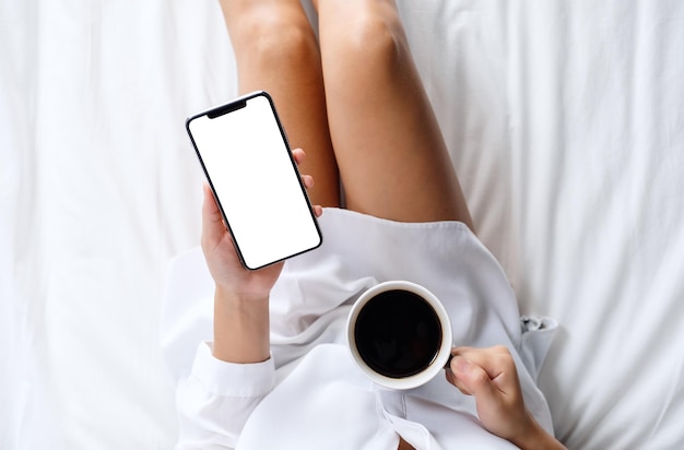 Imagen de maqueta de vista superior de una mujer sosteniendo un teléfono móvil blanco con pantalla de escritorio en blanco mientras toma café en una acogedora cama blanca en casa por la mañana