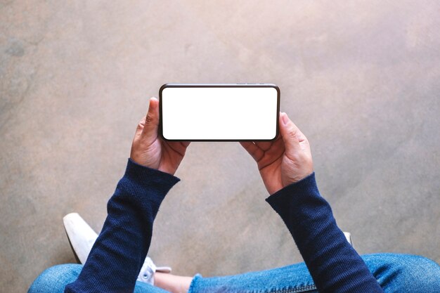 Imagen de maqueta de vista superior de una mujer sosteniendo un teléfono móvil negro con pantalla en blanco mientras está sentada en el suelo