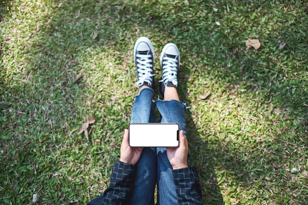 Imagen de maqueta de vista superior de una mujer sosteniendo un teléfono móvil negro con pantalla en blanco mientras está sentado al aire libre