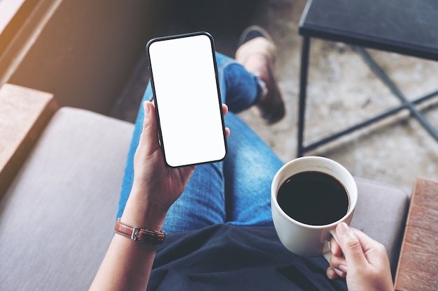 Imagen de maqueta de vista superior de mujer sosteniendo un teléfono móvil negro con pantalla en blanco mientras está sentado y tomando café en la cafetería