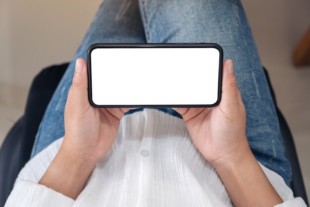 Imagen de maqueta de vista superior de una mujer sosteniendo un teléfono móvil negro con pantalla de escritorio en blanco horizontalmente mientras está sentado