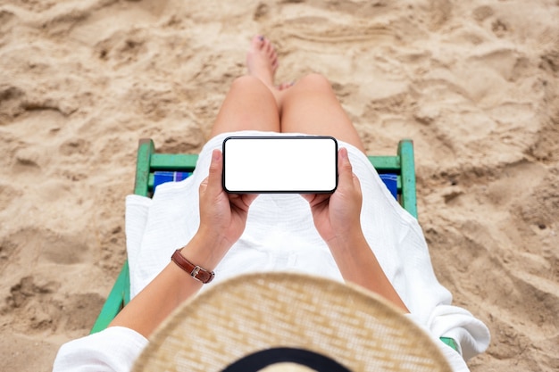 Imagen de maqueta de vista superior de una mujer sosteniendo un teléfono móvil negro con pantalla de escritorio en blanco mientras está acostada en una silla de playa