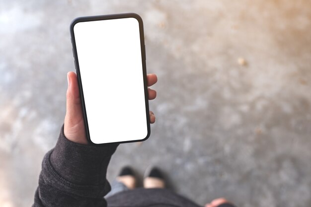 Imagen de maqueta de vista superior de una mujer sosteniendo un teléfono móvil negro con pantalla de escritorio en blanco mientras está de pie en el piso de concreto