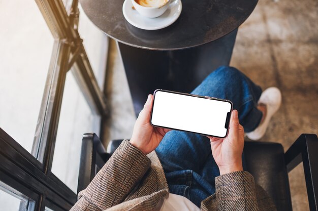 Imagen de maqueta de vista superior de una mujer sosteniendo un teléfono móvil negro con pantalla de escritorio en blanco mientras está sentado en la cafetería