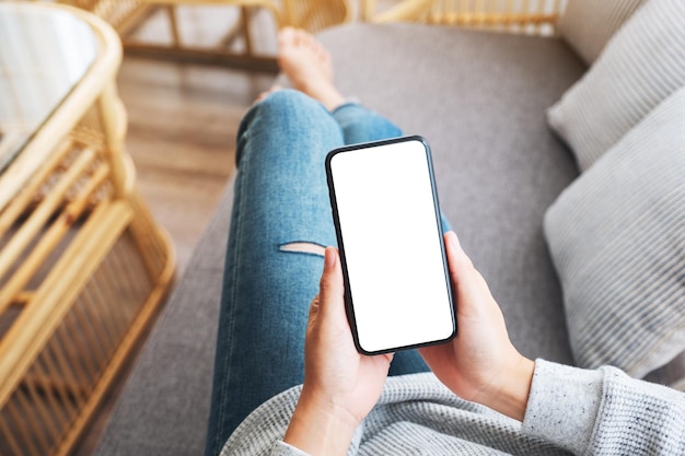 Imagen de maqueta de vista superior de una mujer sosteniendo un teléfono móvil con pantalla blanca de escritorio en blanco mientras está acostada en un sofá en casa