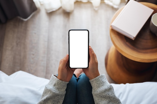 Imagen de maqueta de vista superior de una mujer sosteniendo un teléfono móvil con pantalla de escritorio en blanco mientras está sentada en la habitación de la cama en casa