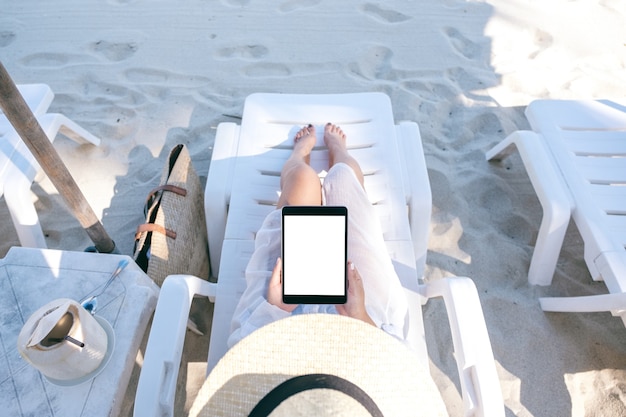 Imagen de maqueta de vista superior de una mujer sosteniendo y usando una tablet pc negra con pantalla de escritorio en blanco mientras se acuesta en una silla de playa en la playa