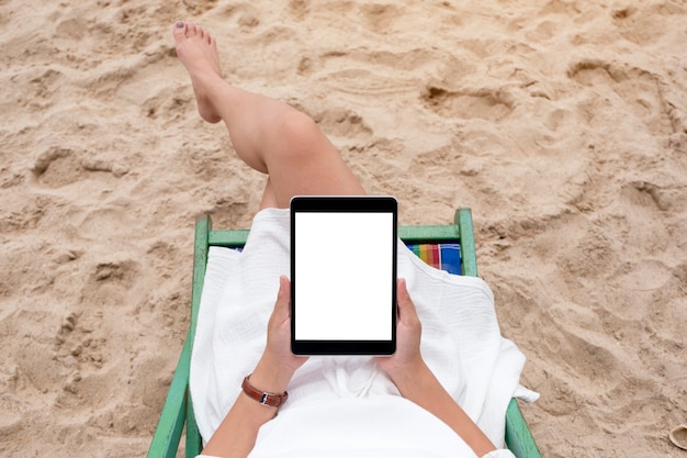 Imagen de maqueta de vista superior de una mujer sosteniendo y usando una tablet pc negra con pantalla de escritorio en blanco mientras está acostada en una silla de playa