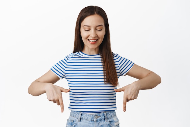 Imagen de una modelo femenina feliz con una camiseta que señala con el dedo hacia abajo, mirando hacia abajo con una cara sonriente y emocionada, mostrando pancartas de venta, publicidad, fondo blanco