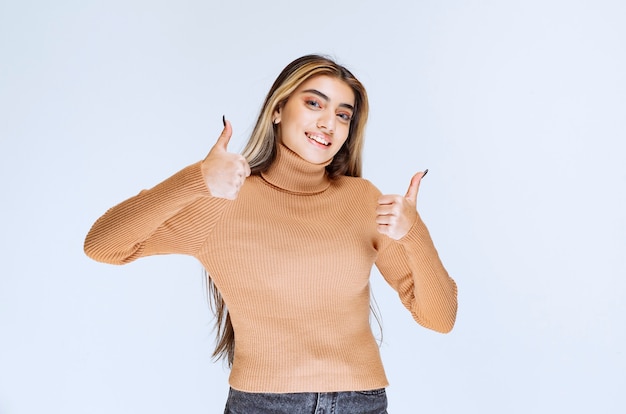 Foto imagen de un modelo de mujer joven en suéter marrón de pie y mostrando los pulgares para arriba.