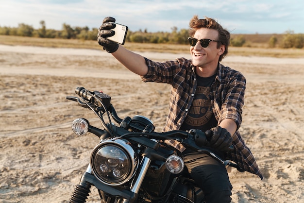 Imagen de motociclista joven optimista feliz guapo en bicicleta al aire libre en el campo del desierto tomar un selfie por teléfono móvil.