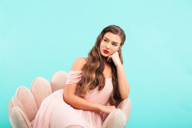 Imagen de mujer aburrida en vestido rosa sentada en un sillón y mirando a un lado con la cabeza apoyada y mirada triste, aislada sobre la pared azul