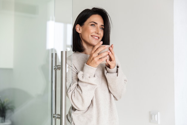 Imagen de una mujer adulta de 30 años sosteniendo una taza con té, mientras está de pie en la moderna habitación luminosa por la mañana