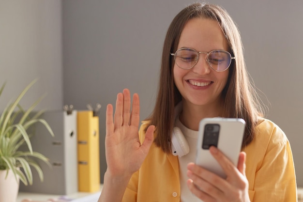 Imagen de una mujer adulta joven de cabello oscuro, alegre y sonriente, independiente usando un teléfono inteligente en la oficina con una videollamada saludando a su amiga reunida en línea