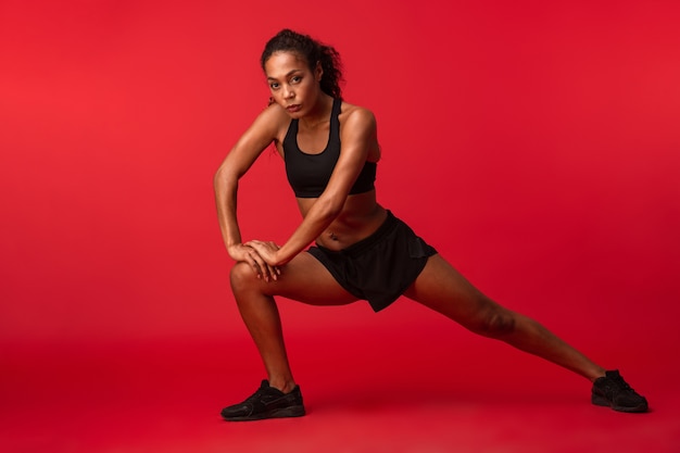 La imagen de una mujer africana joven fuerte de la aptitud de los deportes que presenta aislada sobre la pared roja hace ejercicios.