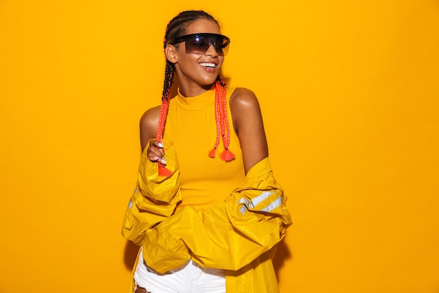 Imagen de mujer afroamericana complacida con gafas de sol y rompevientos sonriendo aislado sobre pared amarilla