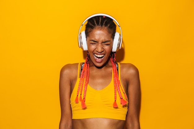 Imagen de mujer afroamericana emocionada con trenzas afro gritando y escuchando música con auriculares aislado sobre pared amarilla