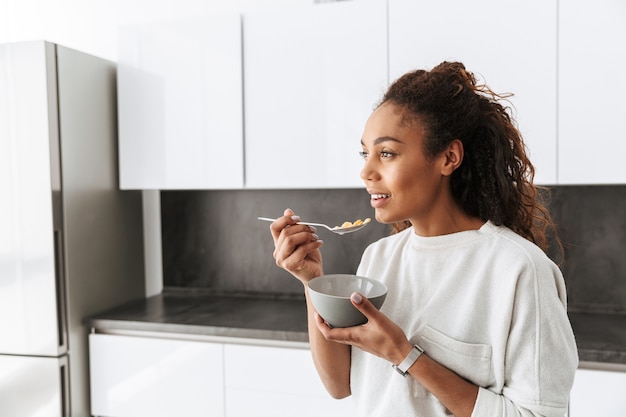 Imagen de mujer afroamericana saludable comiendo copos de maíz con leche en el desayuno, en cocina elegante