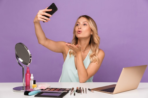 Imagen de una mujer agradable seductora que toma una foto selfie en el teléfono celular mientras envía un beso al aire y se sienta en el escritorio aislado