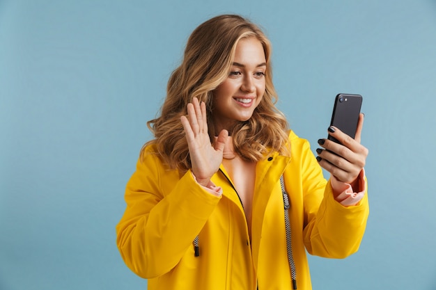 Imagen de mujer alegre de 20 años vistiendo impermeable amarillo sosteniendo teléfono celular y agitando la mano