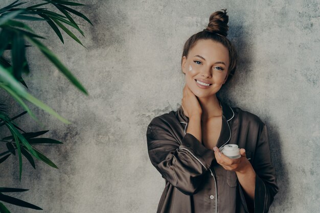 Imagen de mujer alegre en pijama de seda sonriendo mientras se aplica la crema facial, posando con un tarro de loción hidratante en la mano, aislado sobre fondo de muro de hormigón. Concepto de belleza y cuidado de la piel.