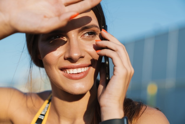 Imagen de mujer alegre en ropa deportiva hablando por teléfono celular mientras está de pie al aire libre en un día soleado