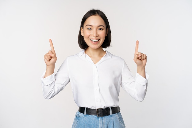 Imagen de una mujer asiática sonriente y segura de sí misma señalando con el dedo hacia arriba mostrando un banner o información de venta de pie sobre fondo blanco