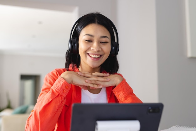 Imagen de una mujer birracial con auriculares sonriendo durante una videollamada en una tableta en casa, espacio para copiar. Trabajar desde casa, tecnología, comunicación y concepto de estilo de vida.